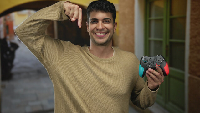 Man grips controller and gestures by pointing amid street bustle where hispanic young outdoor male clutches gamepad while smiling broadly. - Powered by Adobe