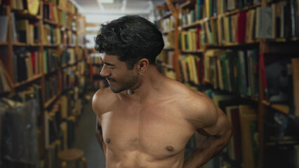 Shirtless young man holds his lower back while standing between shelves in a dimly lit old library aisle; backache discomfort.