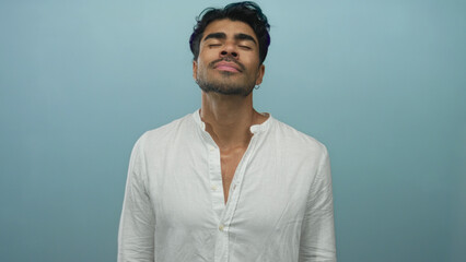 Young hispanic man in white shirt tilts head back and closes eyes revealing face in blue studio; serenity.