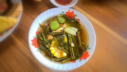 Mixed vegetable tamarind called sayur asem featuring leafy greens, corn, and potato pieces in a rich broth. Cozy, wholesome meal set on a wooden table
