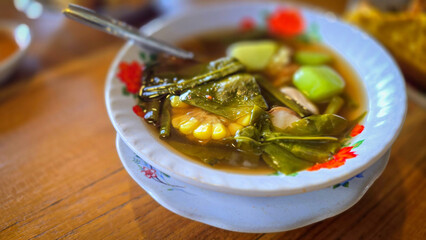 Mixed vegetable tamarind called sayur asem featuring leafy greens, corn, and potato pieces in a rich broth. Cozy, wholesome meal set on a wooden table