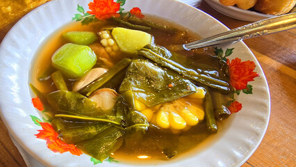 Mixed vegetable tamarind called sayur asem featuring leafy greens, corn, and potato pieces in a rich broth. Cozy, wholesome meal set on a wooden table