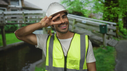 Young hispanic man with hardhat and safety vest flashes peace sign with hand at building entrance; optimism hope positivity confidence.