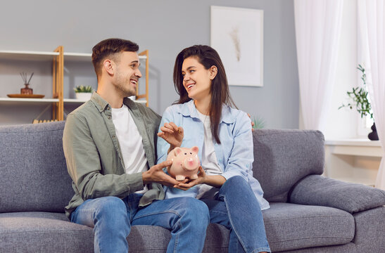 Happy young smiling couple holding piggy bank and putting money coin sitting on sofa at home together. Cheerful man and woman saving money planning budget. Investments and financial freedom concept.