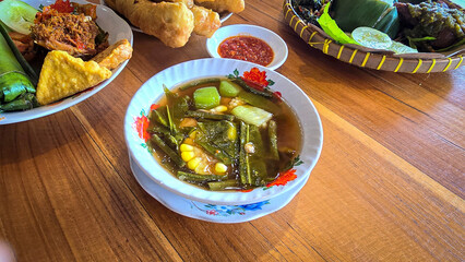 Mixed vegetable tamarind called sayur asem featuring leafy greens, corn, and potato pieces in a rich broth. Cozy, wholesome meal set on a wooden table