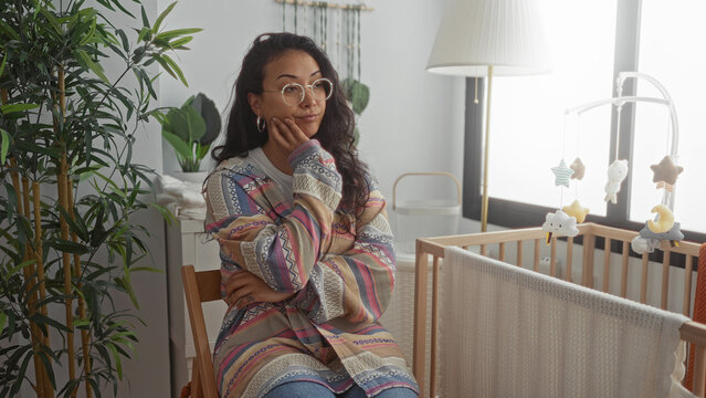 Woman seated by a crib in a building with hand on cheek and colorful cardigan, wearing glasses; motherhood concern.