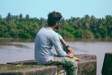 Young man sits by a tranquil river, contemplating the lush palm tree landscape in India. This back view captures a peaceful outdoor moment.