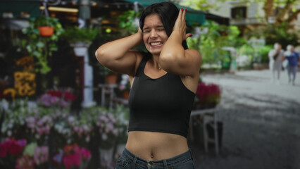Woman covers her ears with her hands in front of a street flower stall under sunlight while...