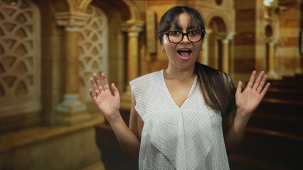 Hispanic brunette woman wearing glasses with hands raised and open mouth singing inside a church...