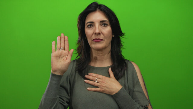 Hispanic middle age woman with raised palm and hand on chest in studio taking an oath gesture; commitment.