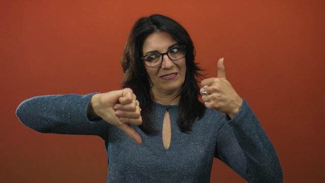 Middle aged hispanic woman smiling and showing thumbs up and thumbs down in studio with orange backdrop; ambivalence.