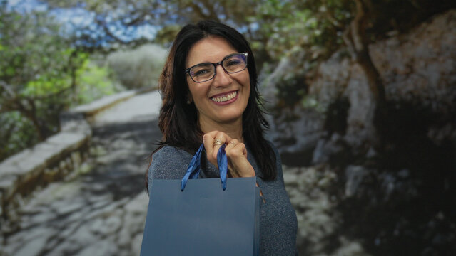 Middle age hispanic woman holds blue shopping bag handles on a sunlit park walkway lined with leafy trees; happiness leisure satisfaction wellness.