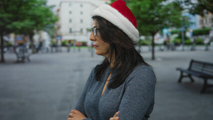 Middle aged hispanic woman wearing santa hat standing with crossed arms and glasses on a street; contemplation.