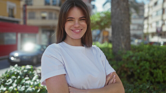 Woman smiling in a city street with arms crossed while wearing a white shirt surrounded by greenery and urban buildings on a sunny day - Powered by Adobe