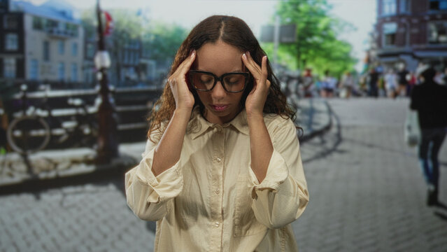 Woman with glasses touching head and hand on chest on urban street by canal and bridge; fatigue contemplation.