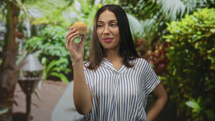 Fototapeta premium Woman holding muffin with raised hand in park amid green foliage and path wearing striped blouse, brunette hair visible; curiosity appetite.