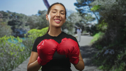Woman holding red boxing gloves up on a forest path, smiling with gloves at chest; confidence training.