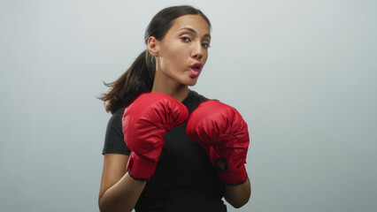 Young hispanic woman wearing red boxing gloves, puckering lips and holding a guard pose in a studio with pale gray wall; playful energy.