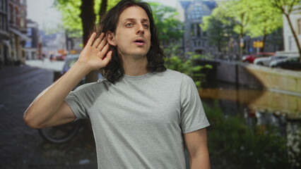 Young hispanic man cupping hand to ear, long hair and gray t shirt, outdoors on a street by a canal in amsterdam; curiosity.