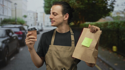 Man holding coffee cup and paper takeaway bag on busy city street with smile and apron; cheerful...