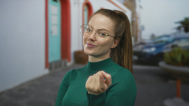 Woman wearing glasses and green top makes beckoning fist gesture on street by building facade; invitation friendly.