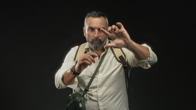Man with backpack and canteen holding up hands, palms forward in studio with black backdrop; adventure determination.