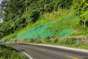 A mountain road with green slope protection and stone retaining wall surrounded by lush forest vegetation
