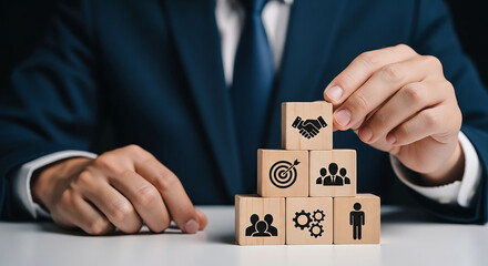 Businessman building a pyramid of wooden blocks with business icons, symbolizing success and teamwork.