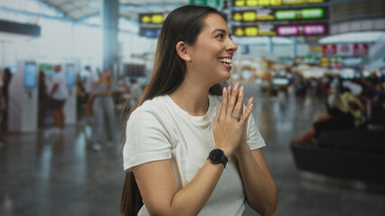 Woman smiling with hands near face touching hair in busy airport terminal, wearing white tshirt and smartwatch; happiness anticipation.