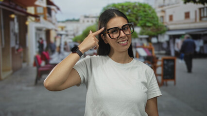 Fototapeta premium Woman points finger to temple at outdoor restaurant terrace on street wearing glasses and white tshirt, smiling slightly; thinking playful.