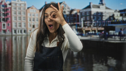 Fototapeta premium Woman makes ok sign around eye while wearing denim apron on amsterdam canal street with historic buildings and water reflections; playful exploration.