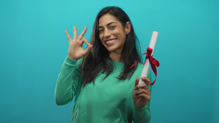 Woman holding rolled diploma tied with red ribbon and making ok sign in turquoise studio; achievement pride success celebration.