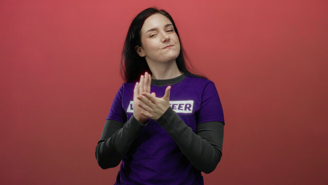 Beautiful brunette woman wearing volunteer shirt claps joyfully in front of vibrant red background radiating happiness and engagement - Powered by Adobe