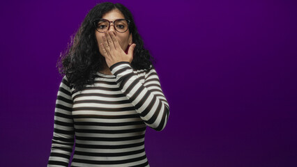 Young arab brunette woman wearing glasses and a striped top with hand covering mouth in studio; shyness.