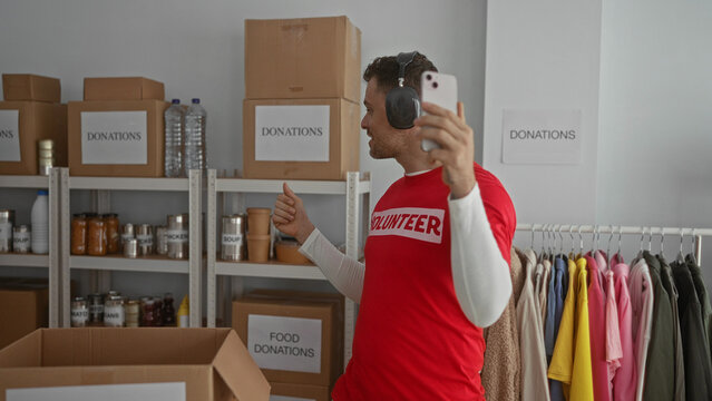 Young man in a volunteer shirt video calling from a donation room surrounded by boxes and clothes, showcasing charity and community engagement indoors.
