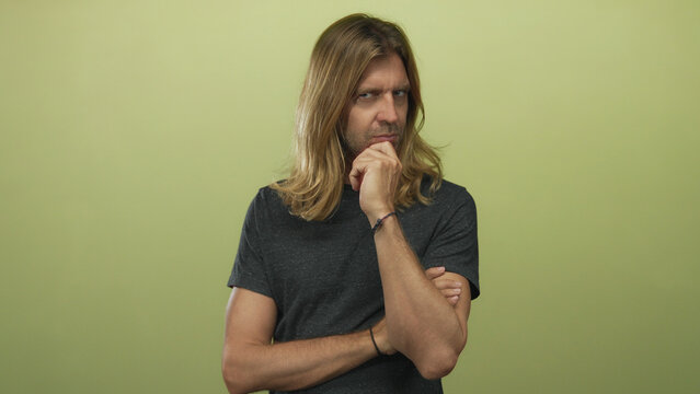Man with long blond hair and hand on chin slightly leaning in studio against a neutral green wall; thinking. - Powered by Adobe
