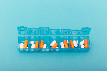 Blue plastic pill organizer with orange and white capsules on turquoise background. The concept symbolizes daily healthcare routine, time management, organization and personal wellness planning