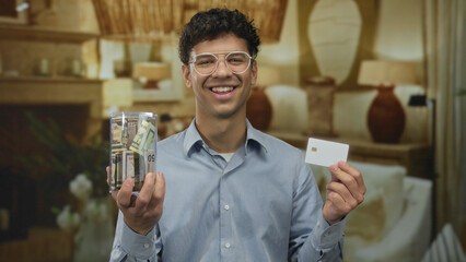 Young hispanic man holds creditcard and jar of rolled banknotes in living room; financial confidence.