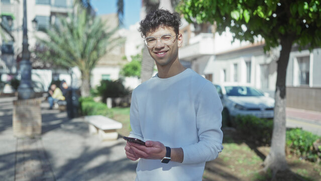 Man tapping smartphone on sunlit street lined with green trees and benches under clear blue sky; connection.