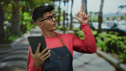 Fototapeta premium Young man in glasses using tablet outdoors in a lush park setting, wearing casual red attire and apron, looking focused and engaging with the touchscreen device.