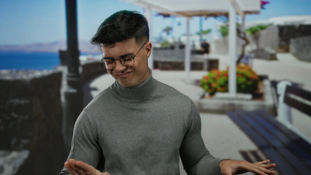 Young man in glasses smiling at seaside promenade with blurred ocean backdrop, wearing gray turtleneck, conveying a casual and relaxed outdoor setting.