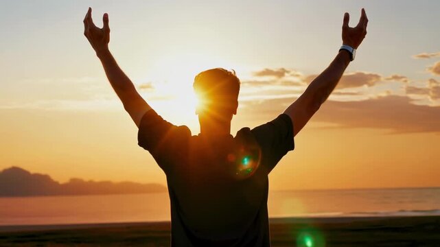 Silhouette of man standing on beach with raised arms facing sunrise over sea expressing freedom success and positive emotional energy in morning light