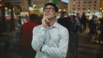 Fototapeta premium Man wearing glasses and light shirt standing with hand on chin gesture in crowded street at dusk under city lights; thinking.