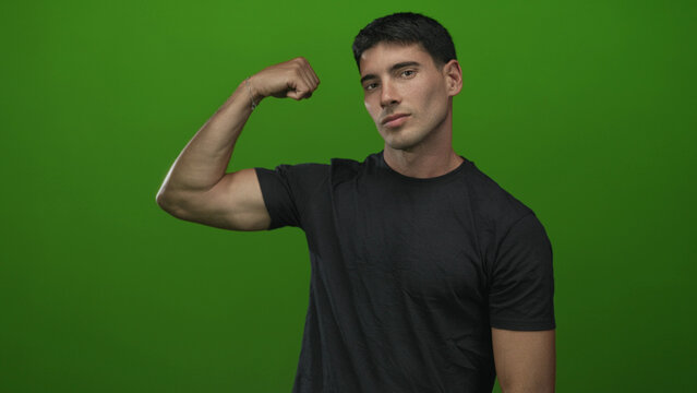 Young hispanic man flexes bicep and inspects muscle in studio against vibrant green backdrop; confidence. - Powered by Adobe