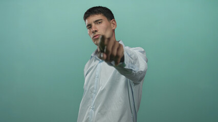 Hispanic man pointing finger to camera with serious gaze in studio wearing light blue shirt;...
