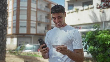 Man holding smartphone and creditcard near building facade on sunny street; digital payment convenience joy.