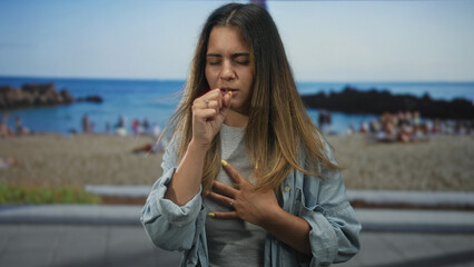 Woman coughing into fist with hand pressed to chest wearing denim shirt and ring at sandy beach...