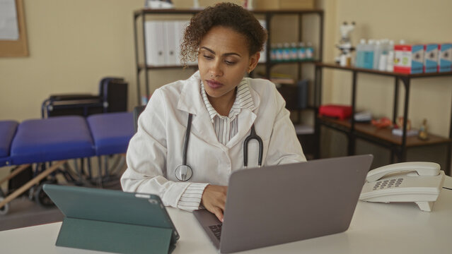 Woman doctor with stethoscope typing on laptop and using tablet at clinic desk in building; concentration compassion care.