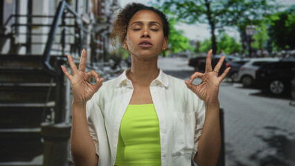 Naklejka premium Woman with closed eyes and raised hands wearing neon green top and white shirt on a street lined with parked cars; calm concentration balance.