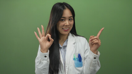 Confident young chinese woman scientist in a lab coat gesturing okay and pointing, isolated against a green wall, suggests scientific excellence and positivity.